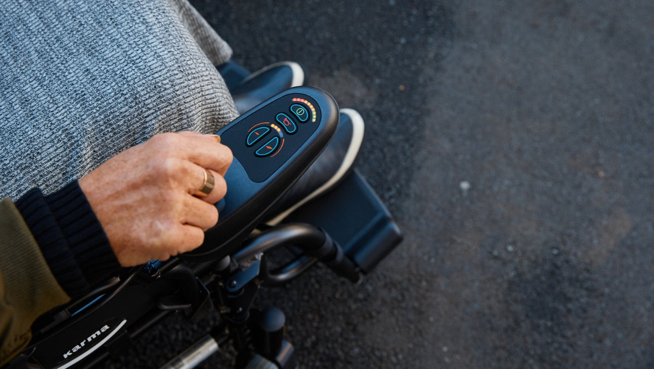 A close-up of a persons hand operating the control panel of an electric wheelchair outdoors. The person has a blanket over their lap.